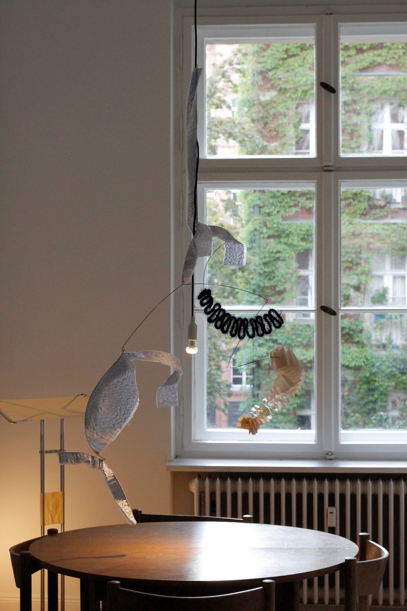 Installation view with a hanging textile mobile by Marina Melentieva, a round table and a window onto a Berlin courtyard